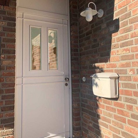 Eine weiße Haustür mit zwei Fenstern, umgeben von einer roten Ziegelmauer, die im Sonnenlicht leuchtet.