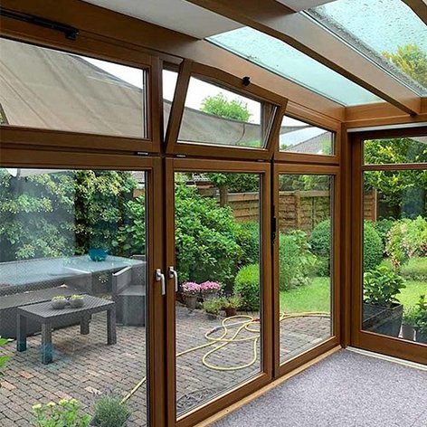 Ein heller Wintergarten mit großen Fenstern, die auf einen gepflegten Garten mit grünen Pflanzen und einem Sitzbereich blicken.