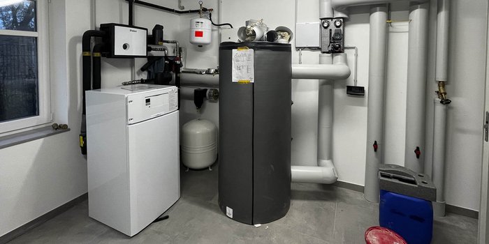 Ein moderner Heizungsraum mit einem weißen Kühlschrank und einem großen, grauen Warmwasserspeicher, umgeben von Rohren.