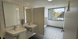 Helle, saubere Sanitäranlagen mit weißen Fliesen, zwei Waschbecken und einem Sichtfenster zur Außenlandschaft.