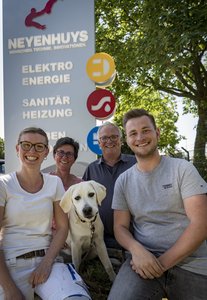 Eine Gruppe von vier Personen und einem Hund posiert fröhlich vor einem Unternehmensschild im Freien.