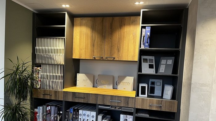 Ein moderner Schreibtisch mit Holzregalen, gefüllt mit Ordnern und Mustern, steht in einem hellen Raum mit Pflanzen.